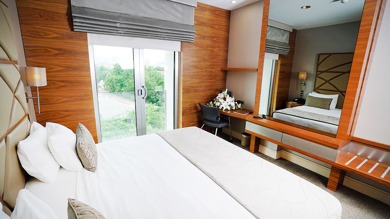 A modern hotel bedroom with a large bed, wooden accents, and a mirrored vanity area.