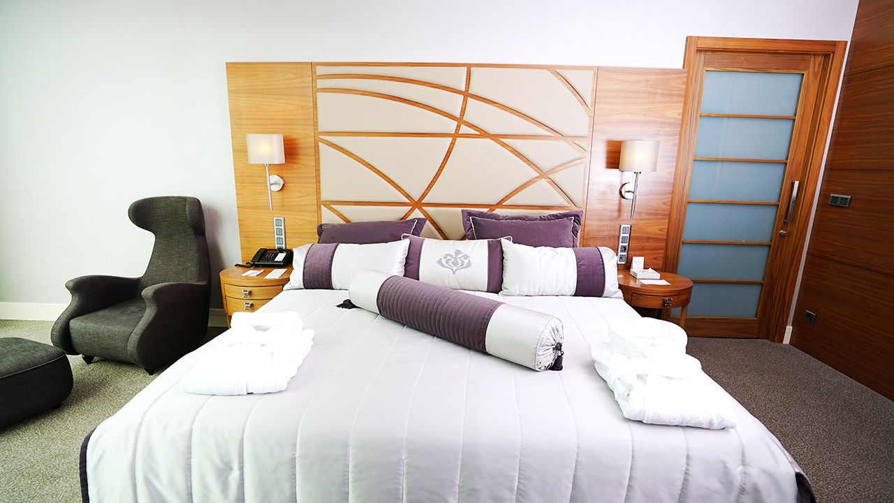Spacious hotel bedroom featuring a bed with purple linens, wood headboard, and armchair.