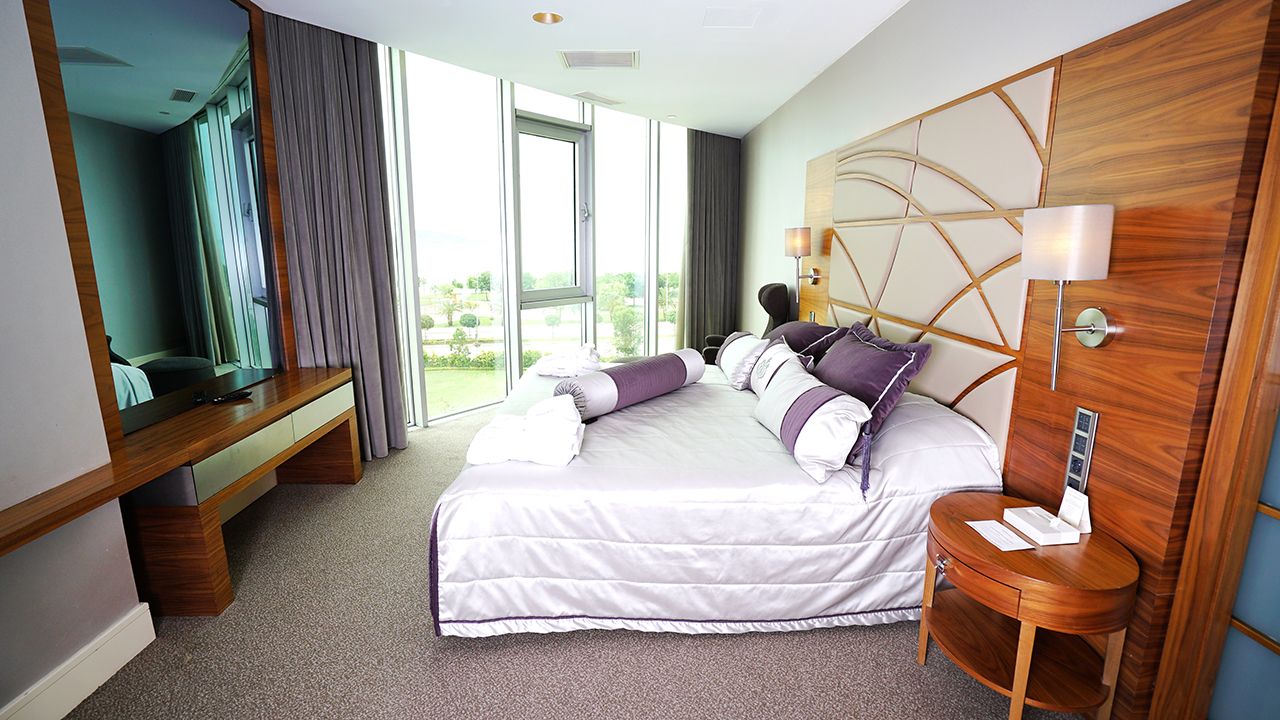 A modern hotel bedroom features a large bed with purple and white pillows, a wooden dresser, and a large window with a view.
