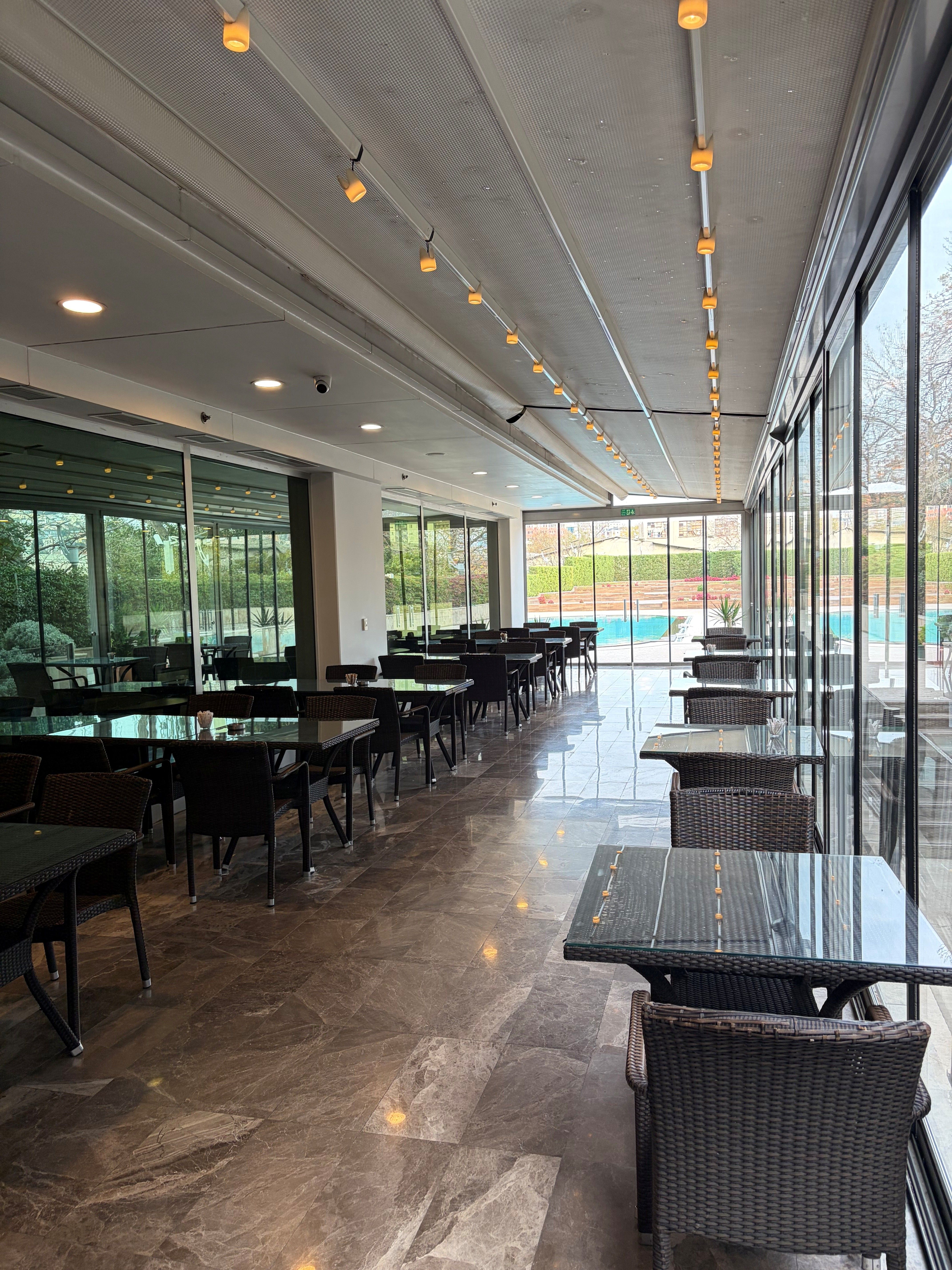 Empty dining area with tables and chairs near a swimming pool visible through large windows.