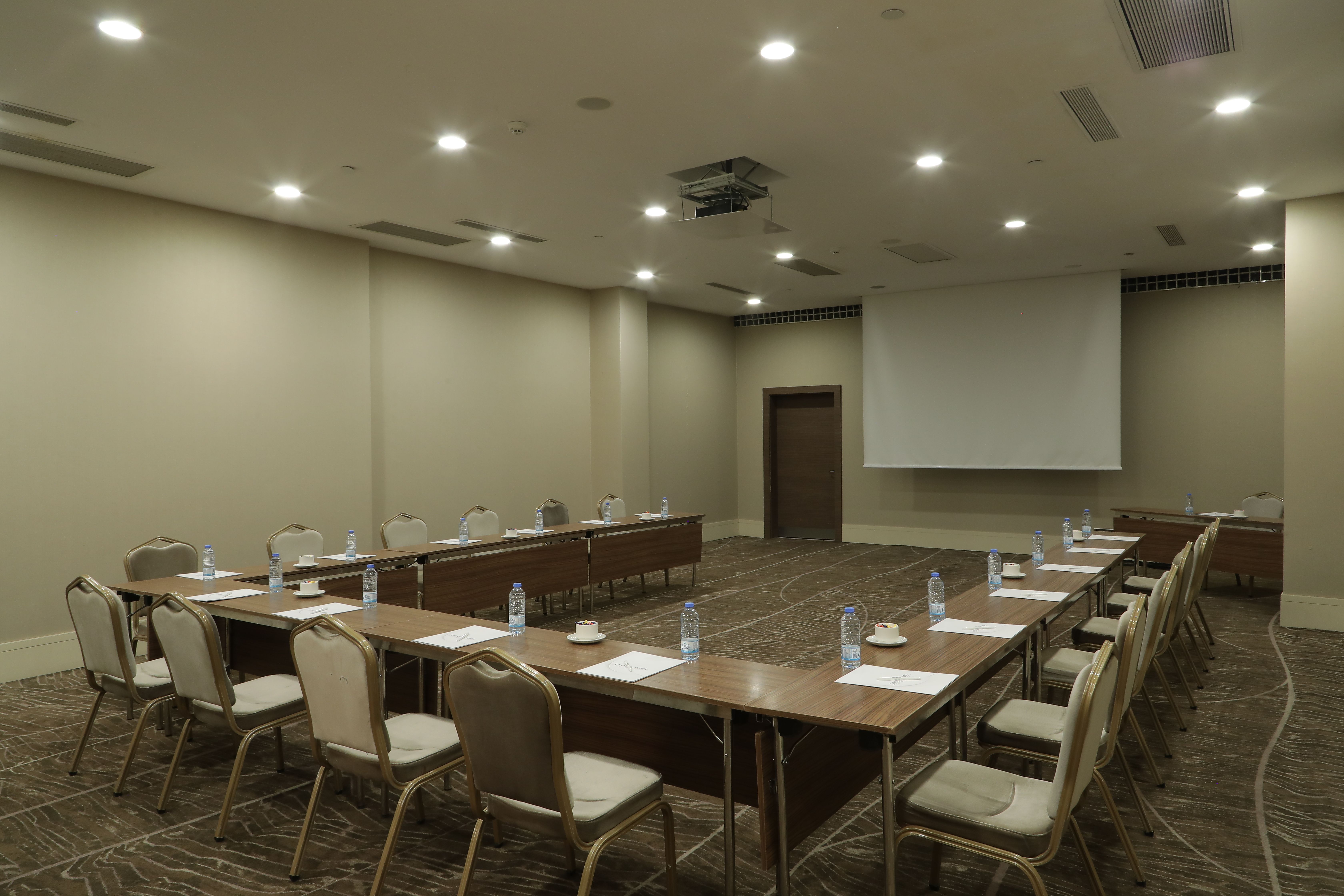 A modern conference room featuring a U-shaped table setup, chairs, and a projector screen.