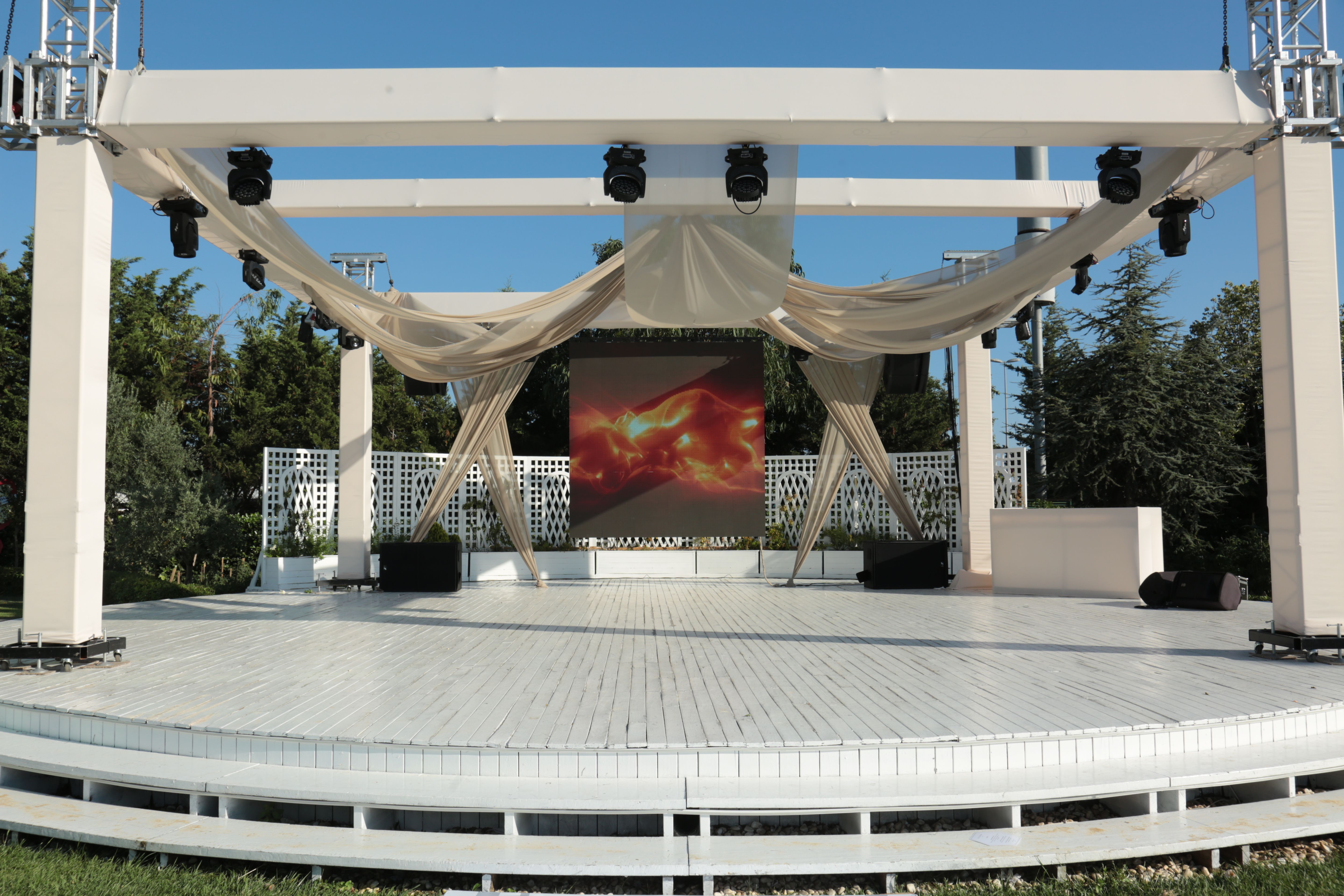 An outdoor stage with a white structure, elegant drapes, and a large screen displaying abstract art.