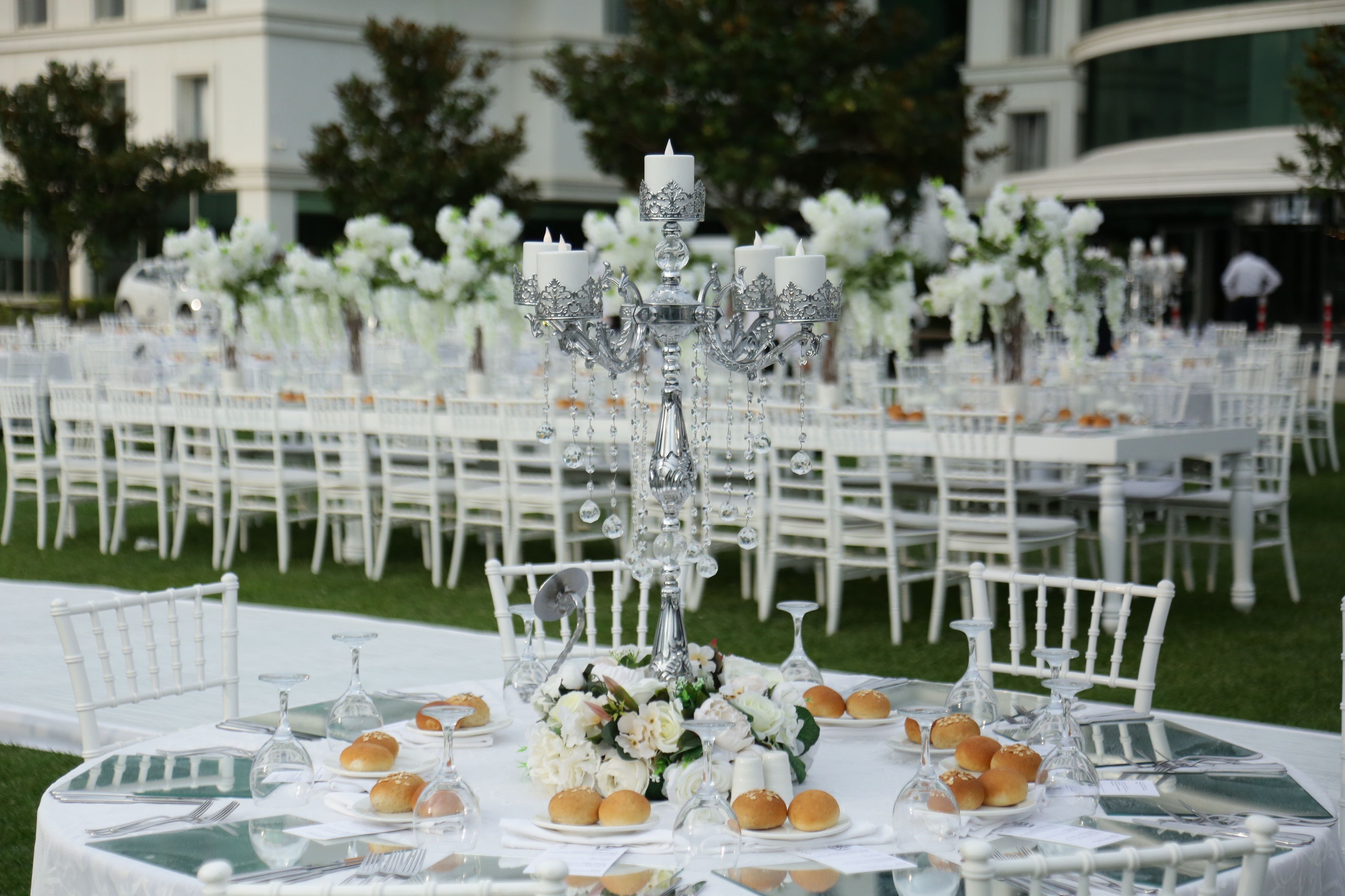 A long, white banquet table decorated with white flowers and candles is set up outdoors on a green lawn.