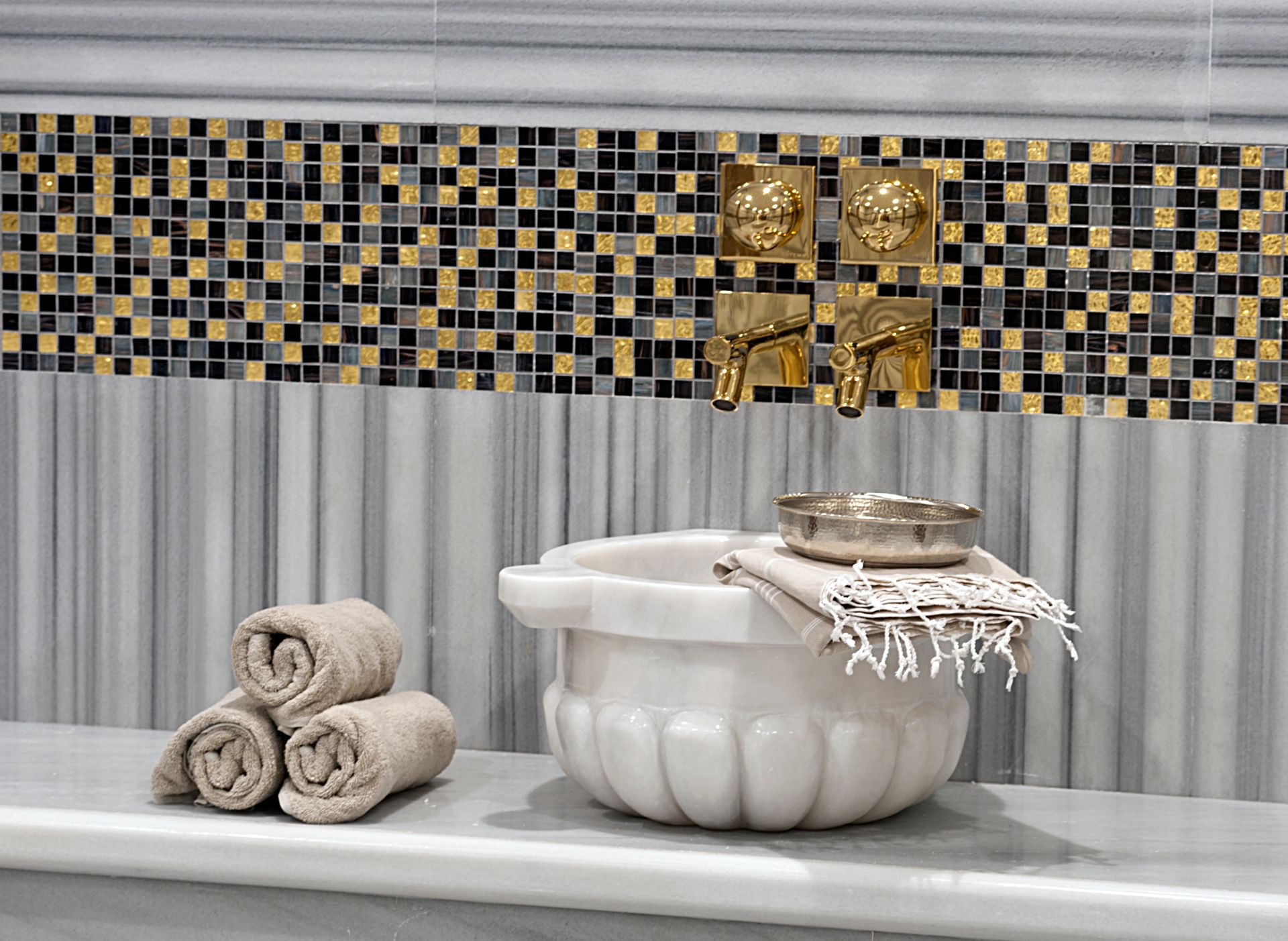Rolled beige towels sit next to a white marble basin with ornate gold fixtures above.