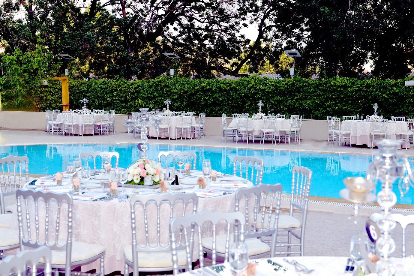 Elegant outdoor event with beautifully set tables surrounding a clear blue swimming pool.