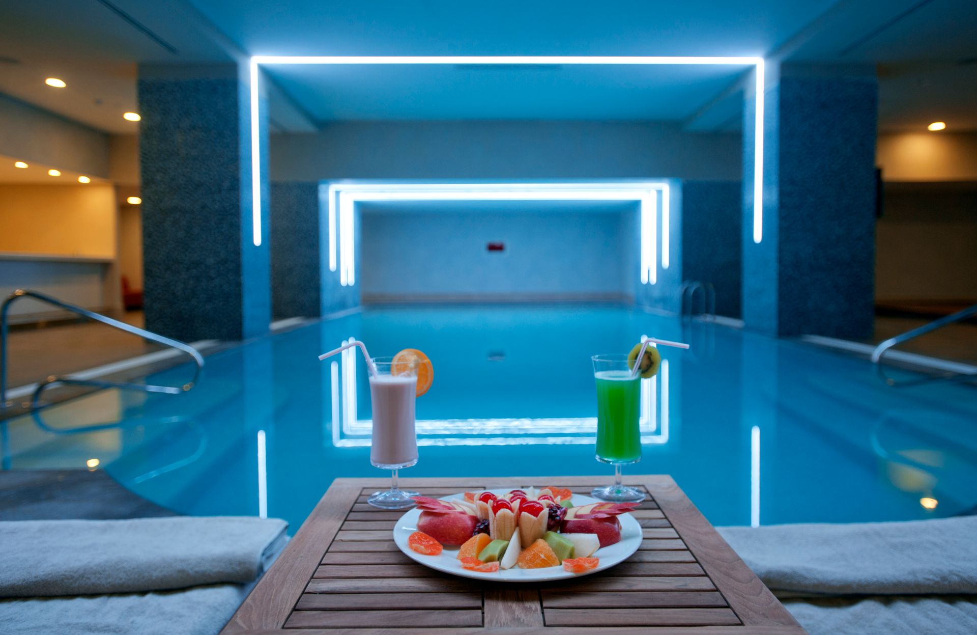 A plate of fruit and two colorful cocktails sit on a table beside an indoor swimming pool with blue neon lighting.