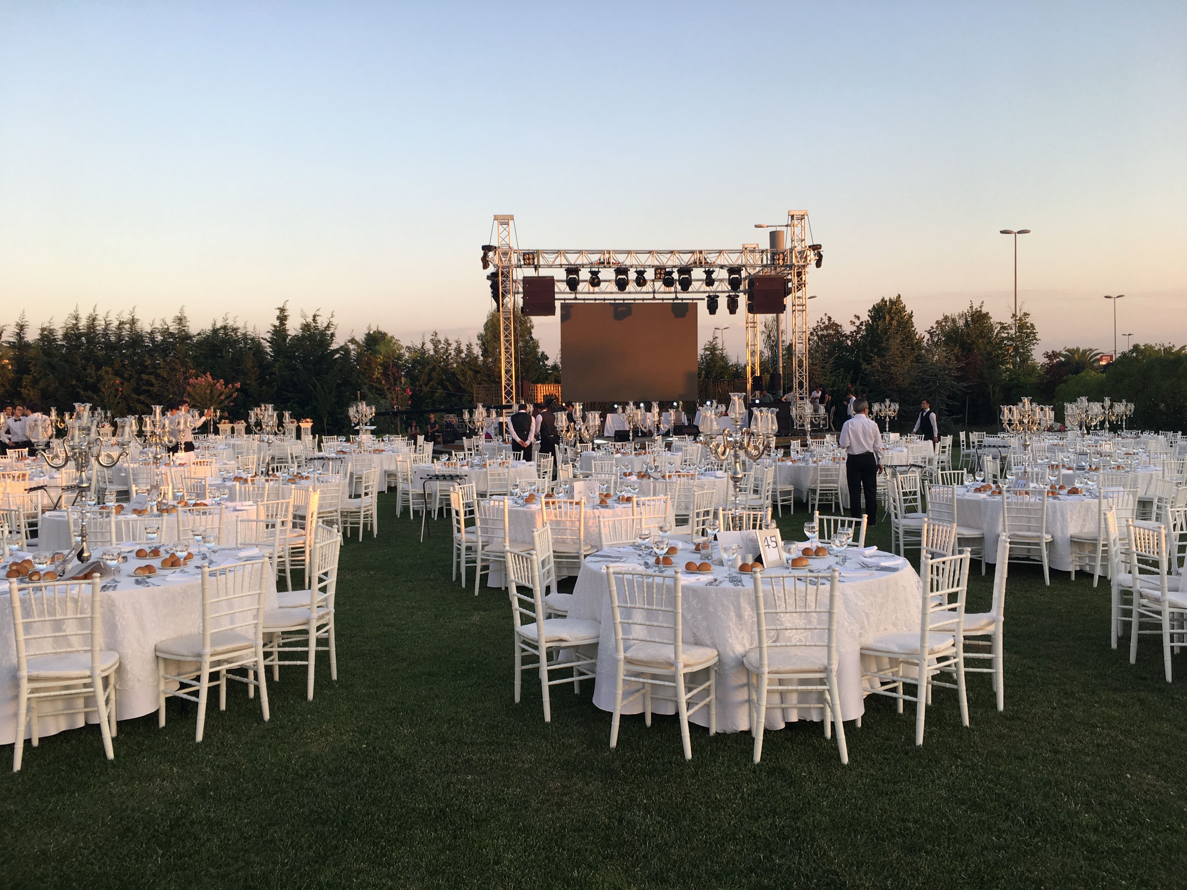 Outdoor event space with white tables and chairs on grass, stage in background.