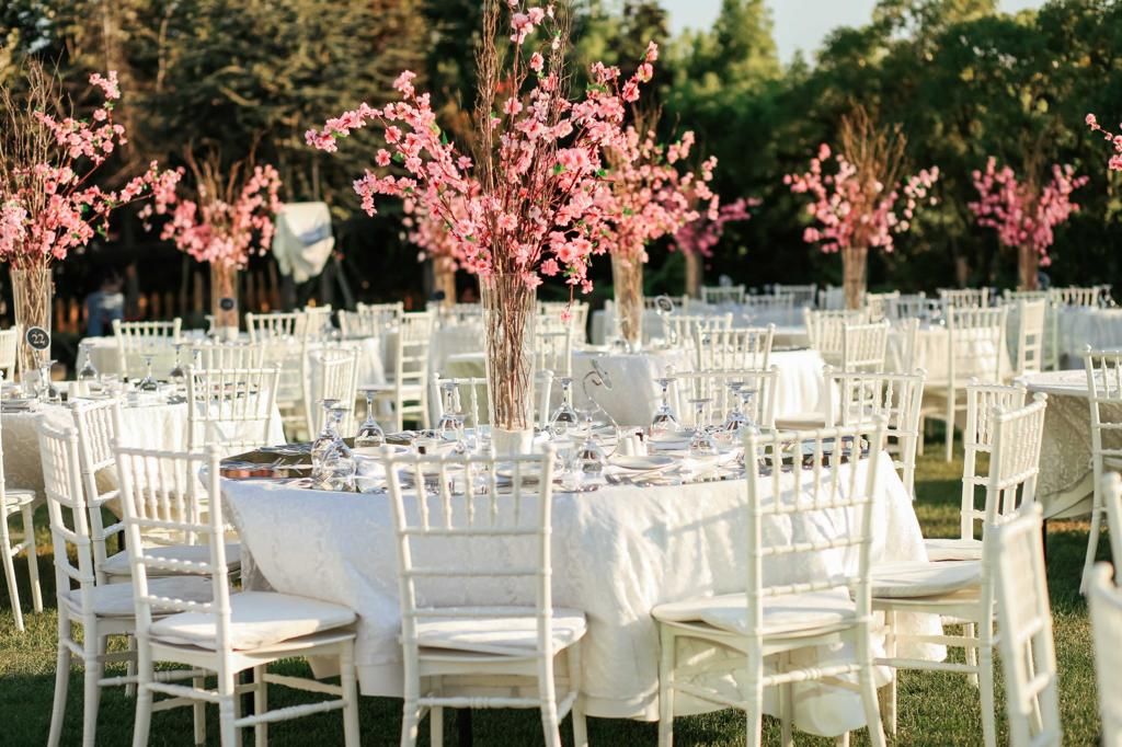 Outdoor event with white tables, chairs, and pink flowering tree centerpieces in a garden.
