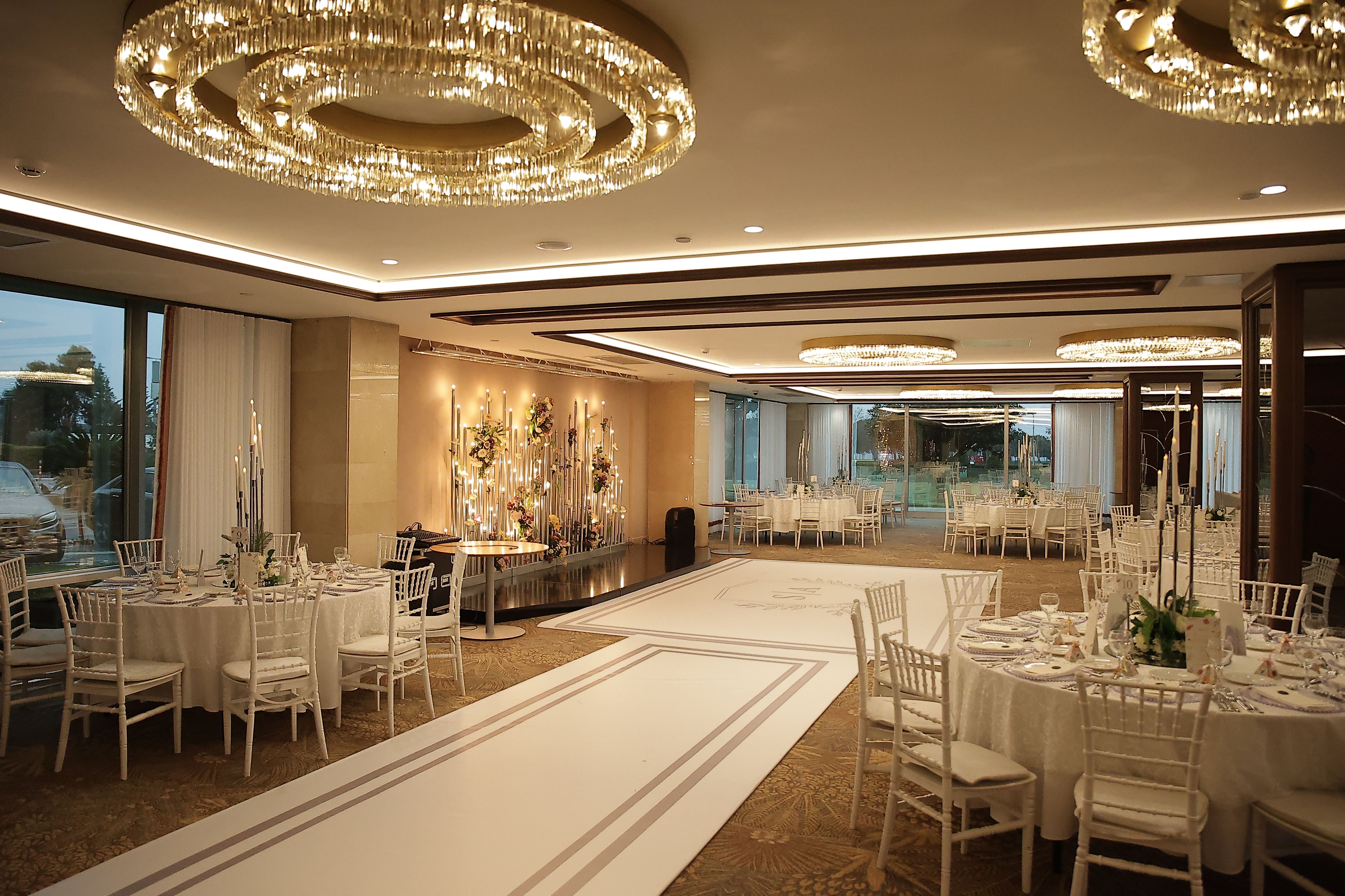 An elegant ballroom features round tables, white chairs, and large chandeliers, set for a formal event.