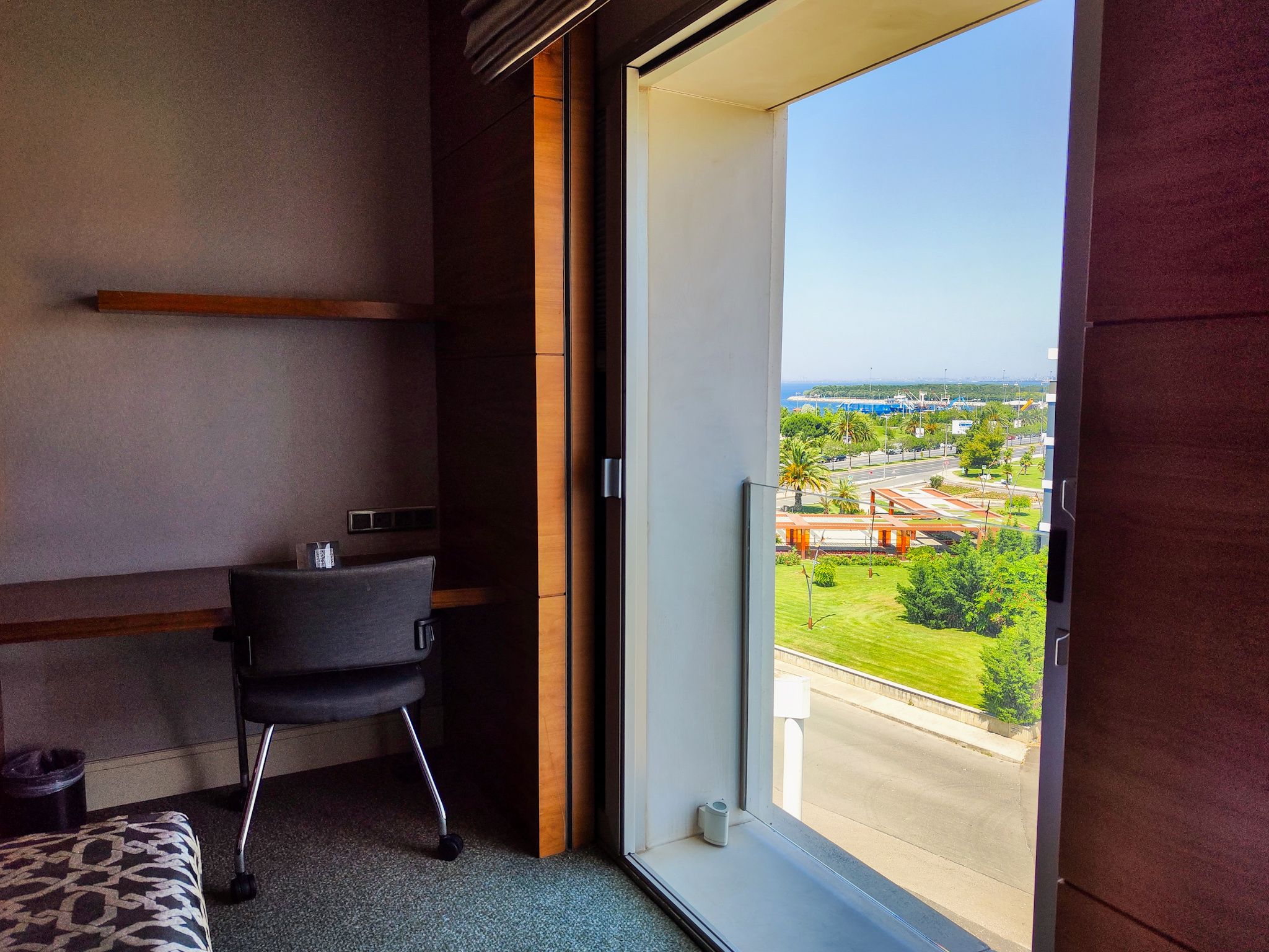 A room with a desk and chair, large window shows bright city vista.