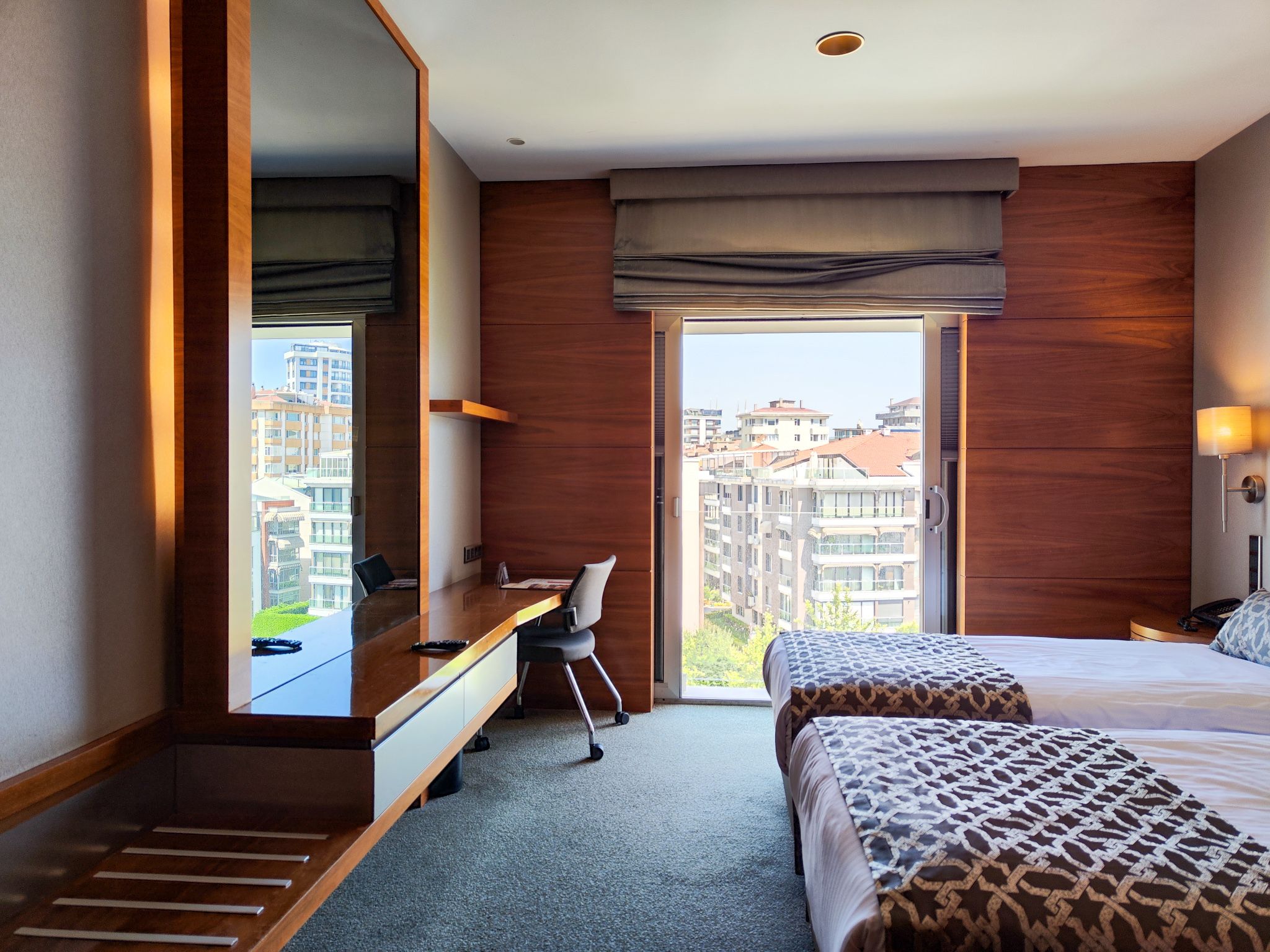 Modern hotel room with two twin beds, a desk area, and a window overlooking a city.