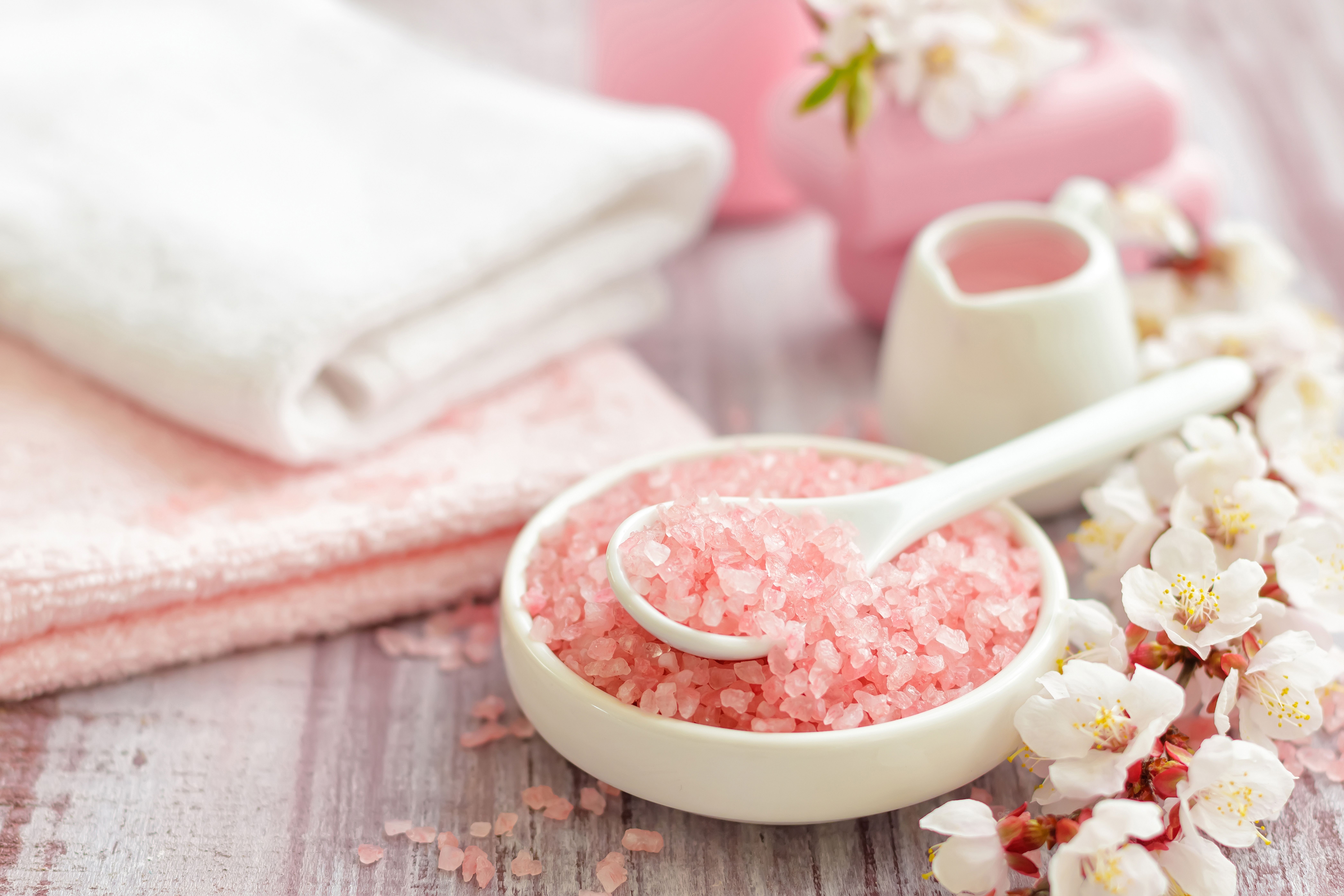 A bowl of pink bath salts with a white spoon rests on a wooden surface with fluffy white and pink towels and delicate white blossoms.
