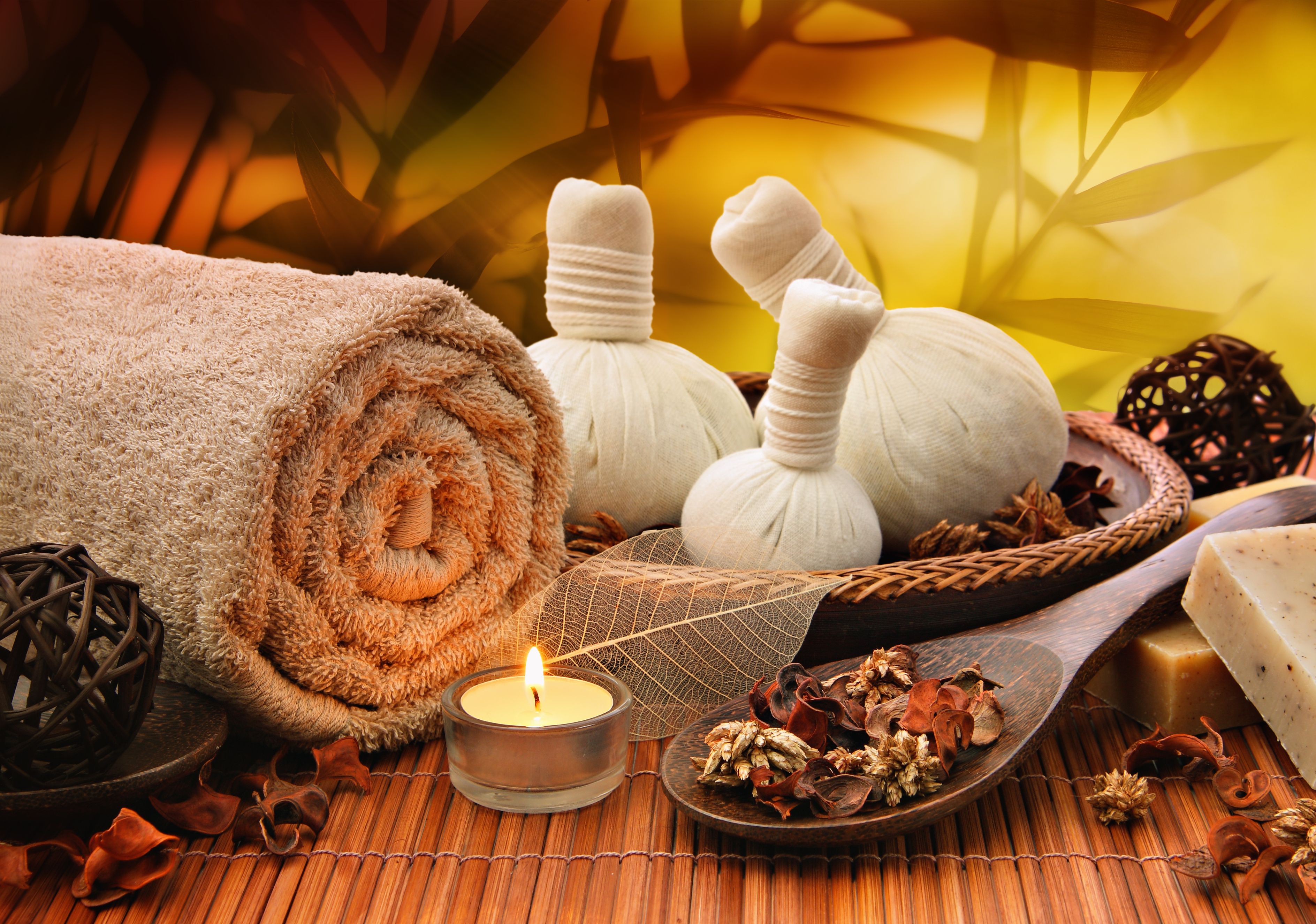 Rolled towel, herbal poultices, candle, and spa ingredients arranged on a wooden surface with bamboo background.