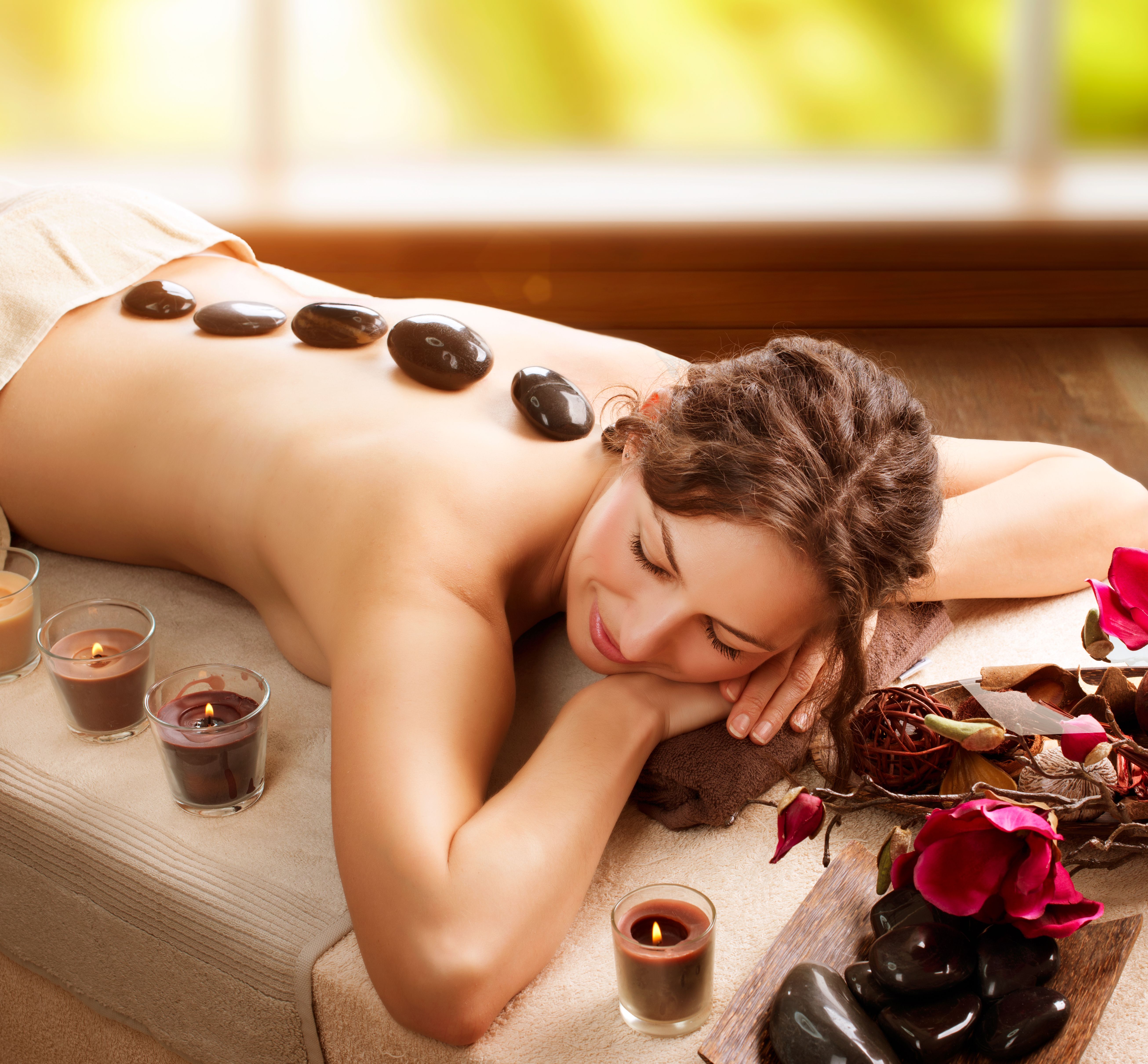 A woman receives a hot stone massage on her back with candles and rose petals nearby.
