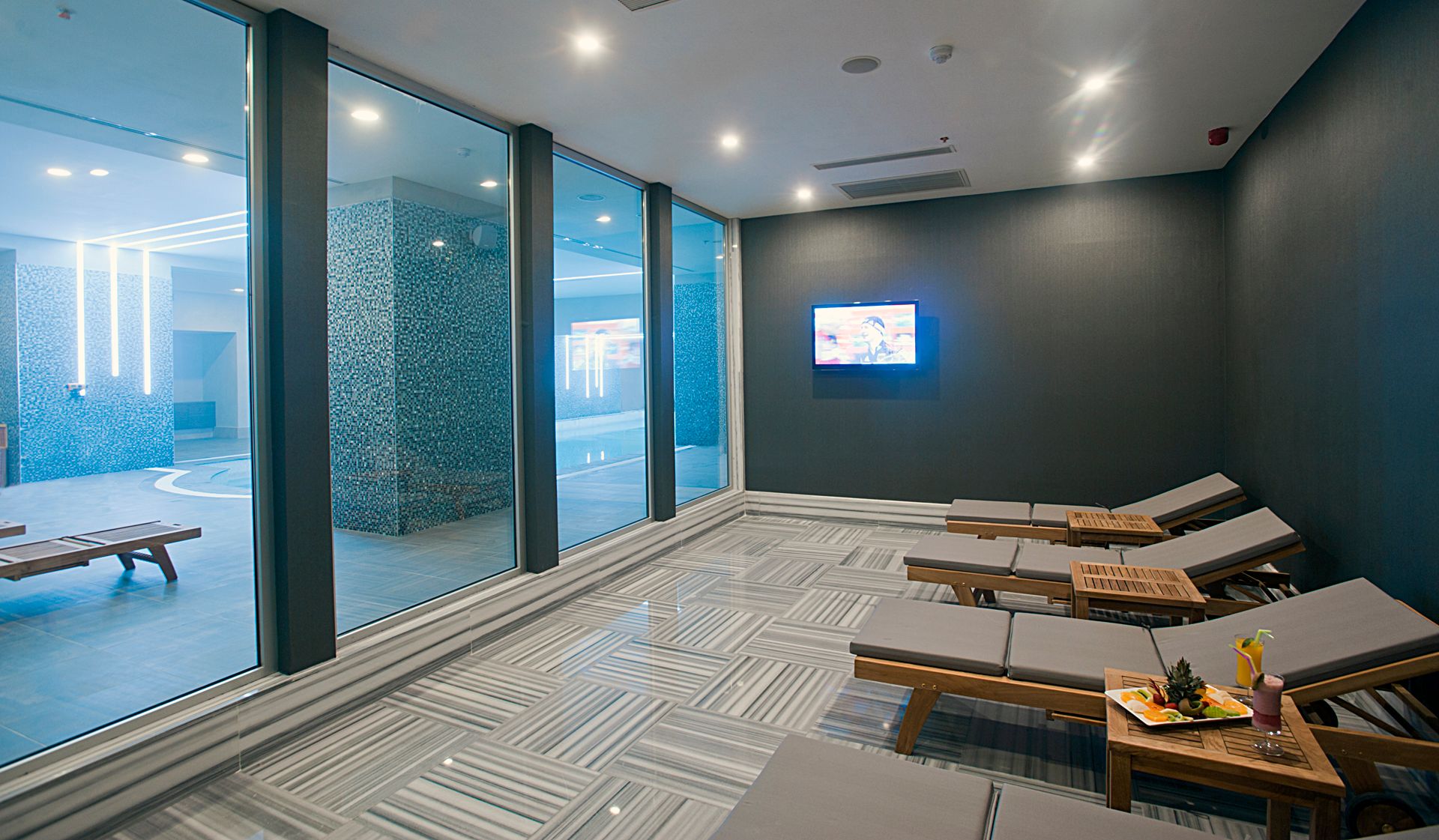 Modern spa relaxation area with chaise lounges and large windows overlooking a pool.