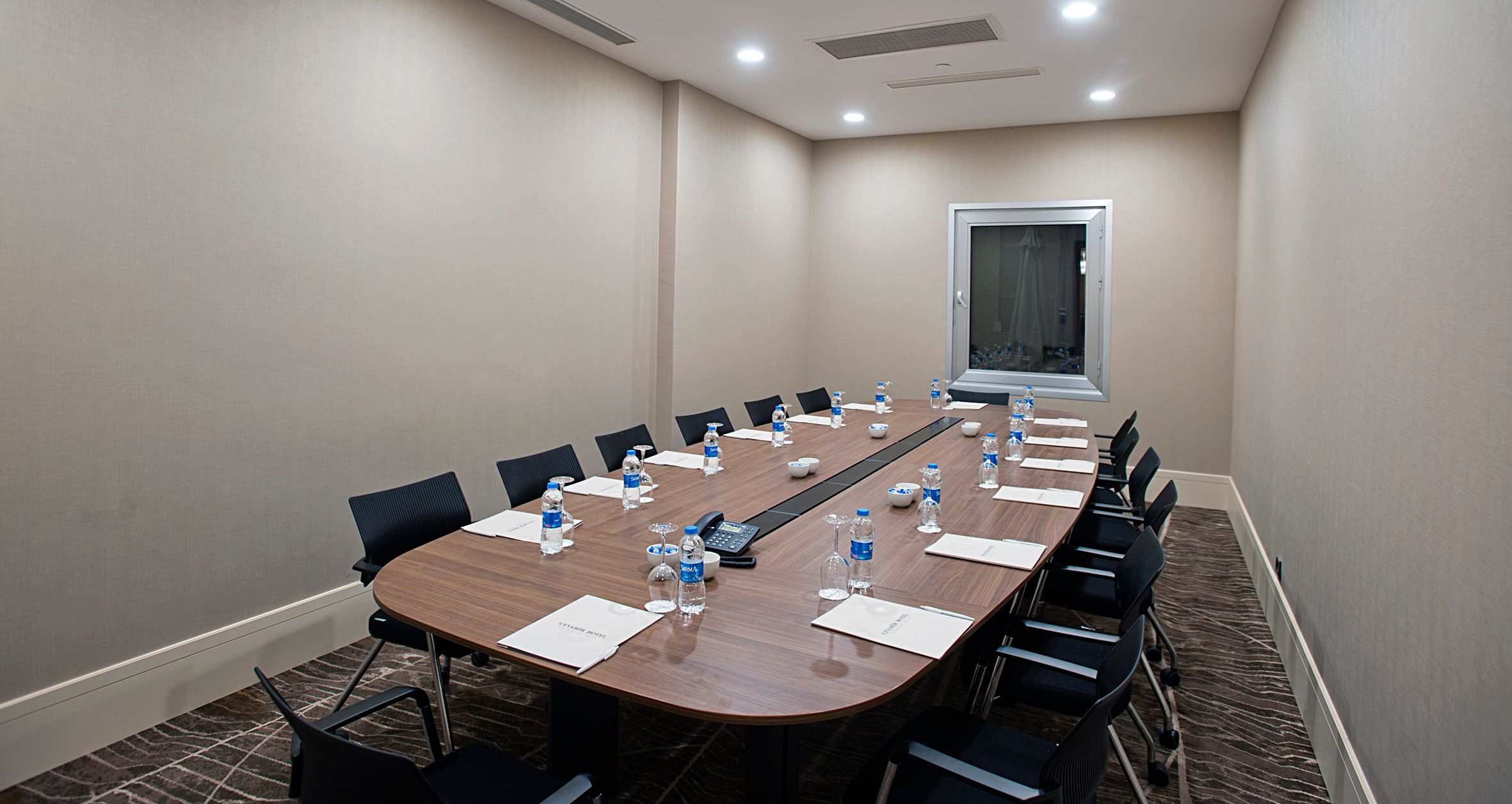 Empty modern meeting room with an oval wooden table, chairs, water bottles, and papers.
